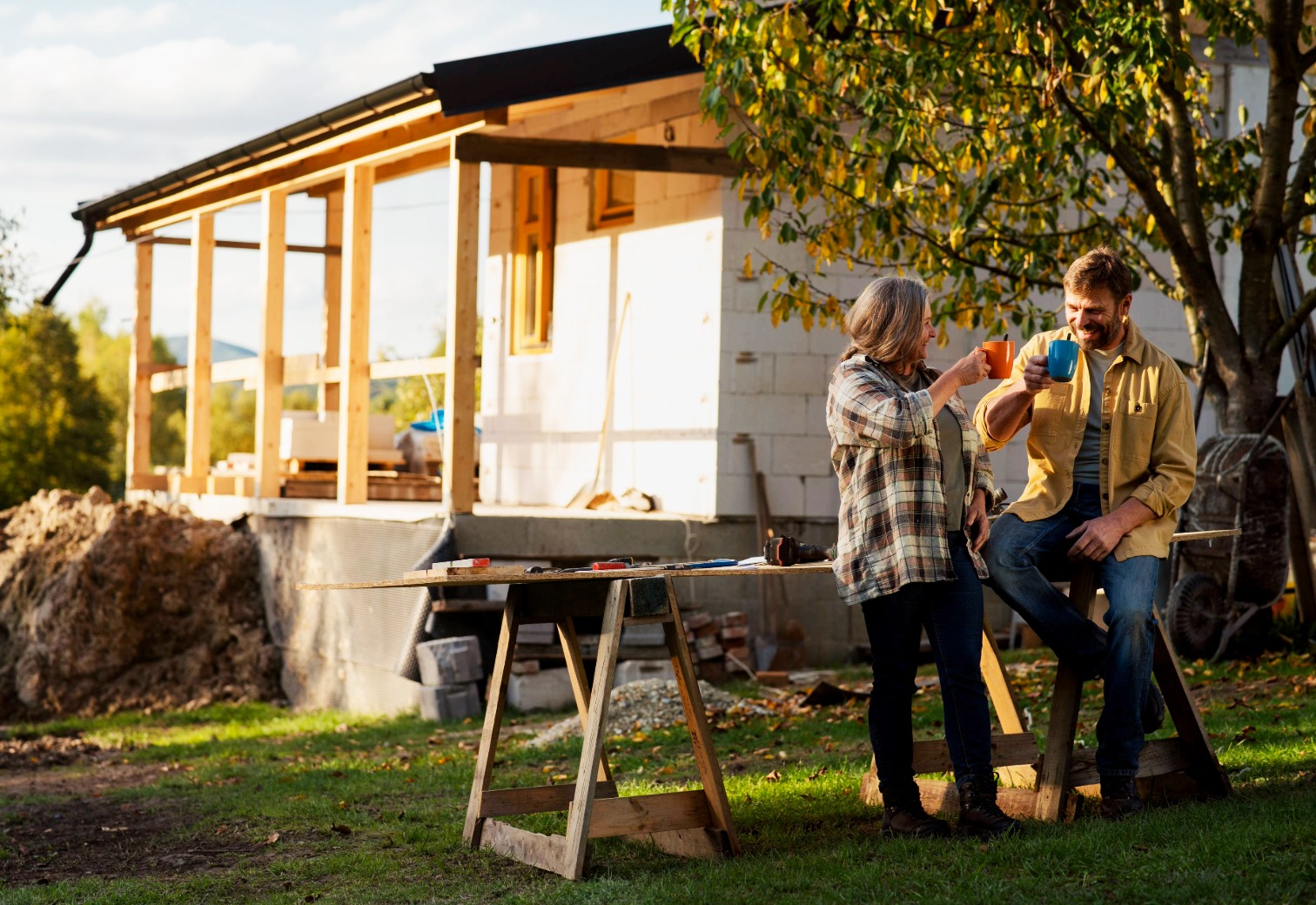 A couple raising coffee mugs beside a sawhorse work table in front of their home mid-renovation